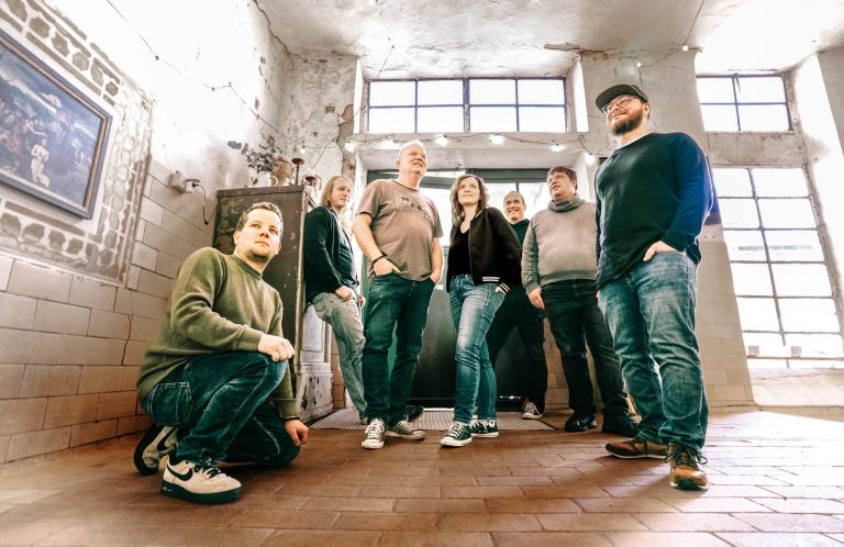 Streugut Sechs Personen stehen in einem Raum mit großen Fenstern und Backsteinwänden.