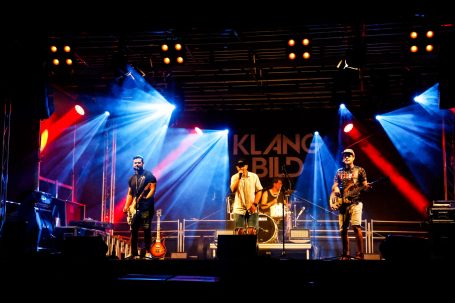 Eine Band spielt auf einer Bühne mit bunten Lichtern und einer Publikumskulisse.