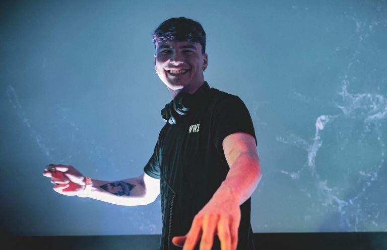 Junger DJ in schwarzem T-Shirt, lächelt und hebt die Hand vor einem bunten Hintergrund.