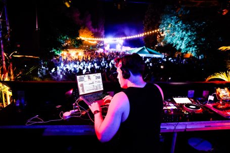 DJ steht an einem Mischpult, spielt Musik für eine feiernde Menschenmenge im Freien.