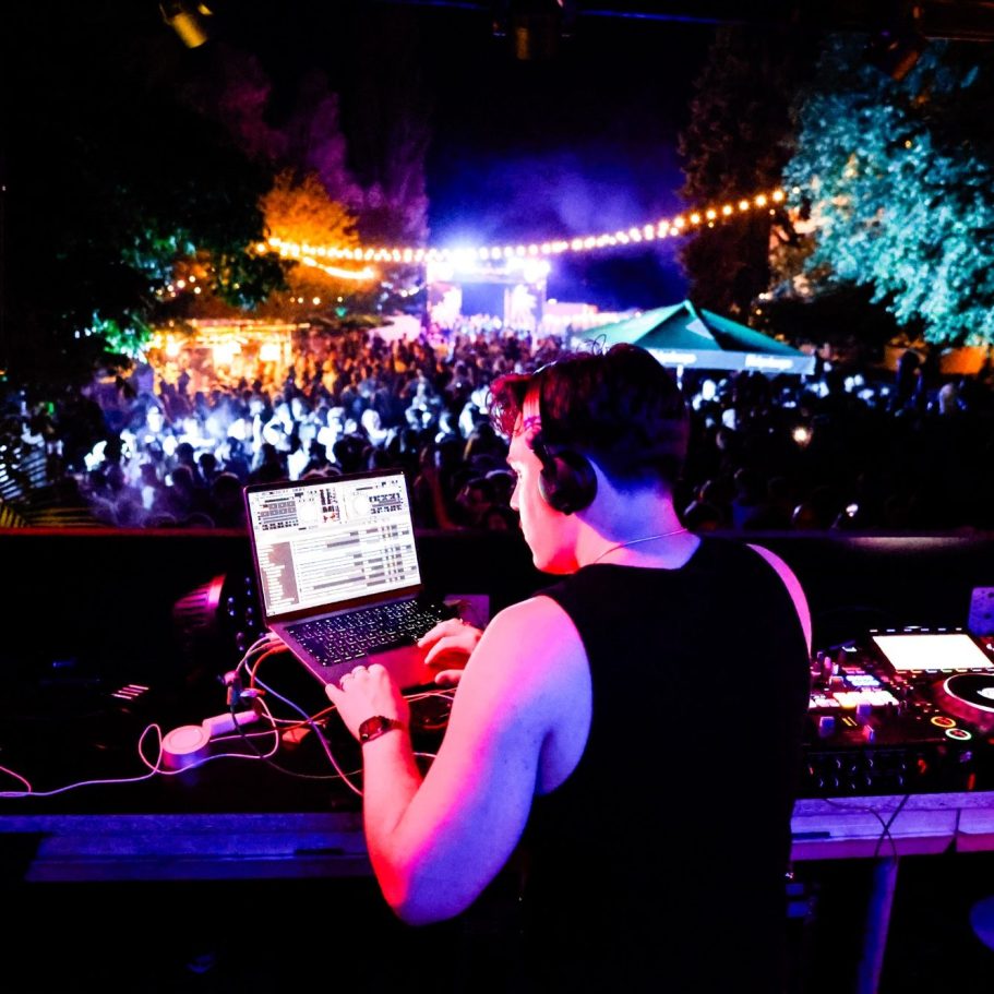 DJ-Pult mit Laptop und Musik-Equipment, Blick auf tanzende Menge im Freien.