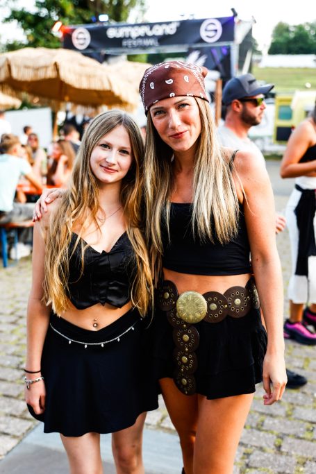 Zwei Frauen in schwarzen Outfits und Accessoires, lächelnd auf einem Festival.