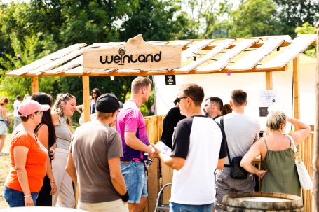 Menschen stehen an einem Stand mit einem Schild "Wajuhund" in einer begrünten Umgebung.