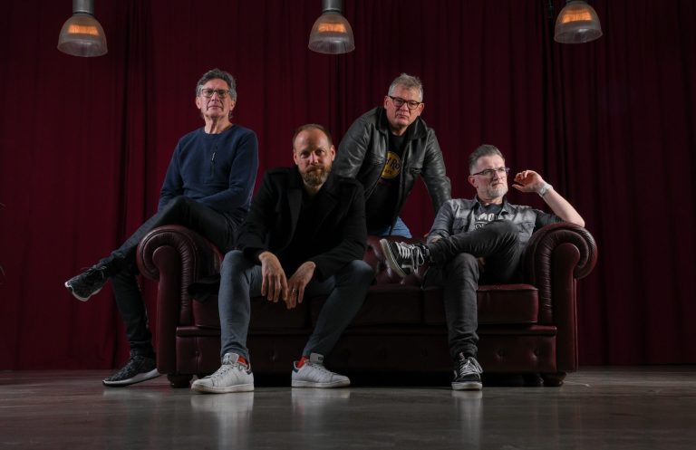 SPALPEENZ Band Vier Männer sitzen auf einer Couch vor einem roten Vorhang, lässig gekleidet.