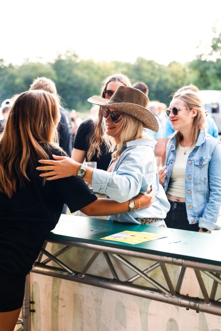 Eine Person mit einem Hut umarmt eine andere an einem Veranstaltungstisch, im Hintergrund stehen weitere Menschen.