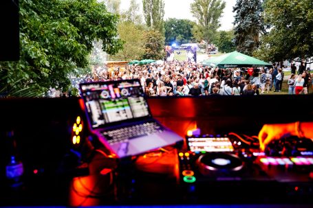 DJ-Pult mit Laptop und Mischpult, Blick auf eine Menschenmenge und Festzelte im Hintergrund.