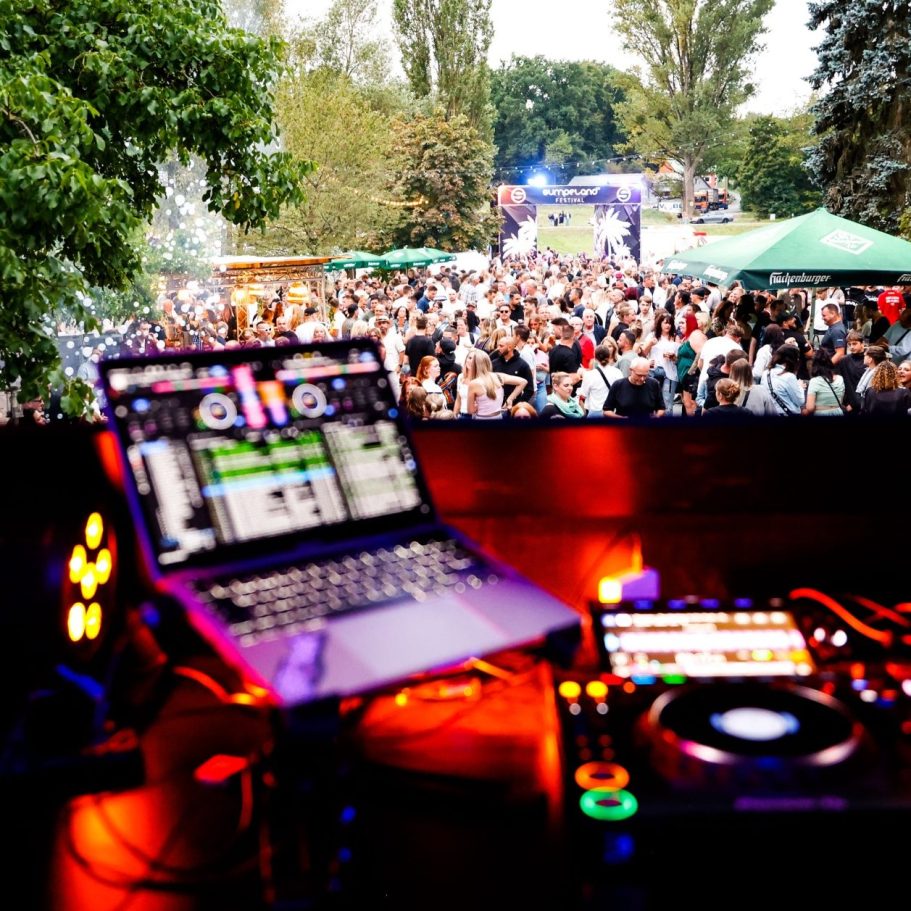 DJ-Pult mit Laptop und Musik-Equipment, Blick auf tanzende Menge im Freien.