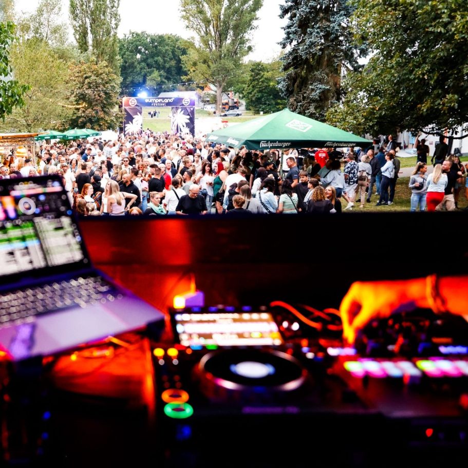 DJ-Pult mit Laptop; Menschenmenge bei einem Open-Air-Event im Hintergrund.