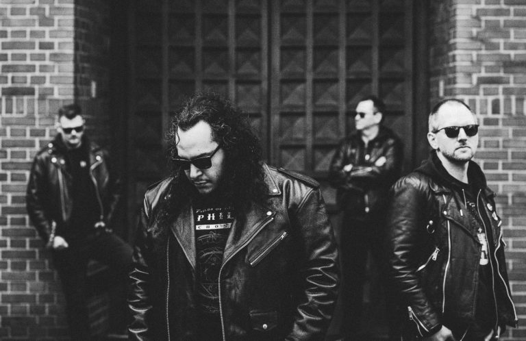 Vier Männer in Lederjacken posieren vor einer Backsteinmauer in Schwarzweiß.