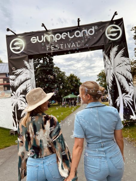 Zwei Frauen halten Händchen und betreten ein Festival mit Palmen und einem großen Banner.
