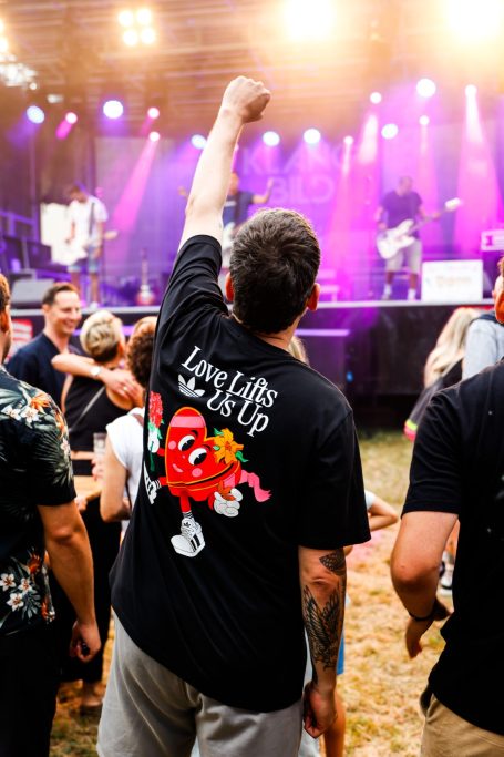 Junger Mann mit gestrecktem Arm vor einer Konzertbühne, trägt ein schwarzes T-Shirt.