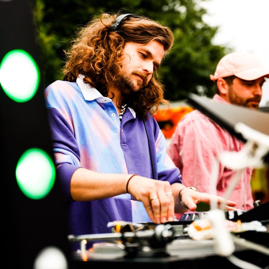 Ein DJ mit lockigem Haar mixt Musik an einem Plattenspieler inmitten von Menschen.