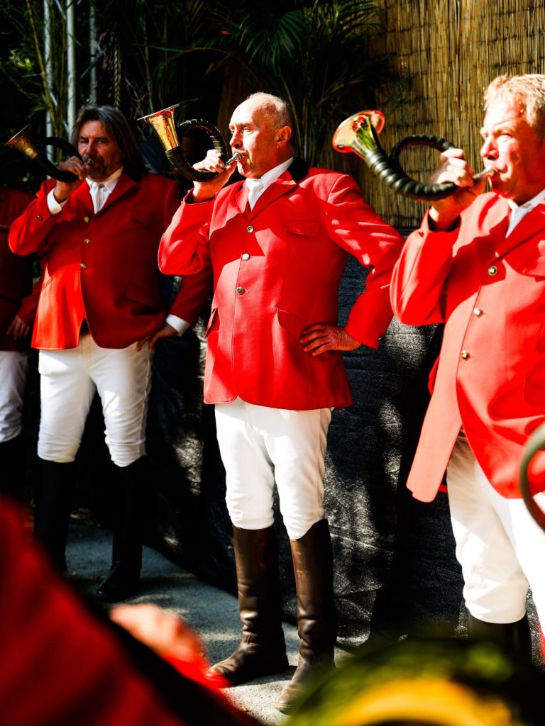 Drei Männer in roten Jacken blasen Blasinstrumente bei einer Feier.