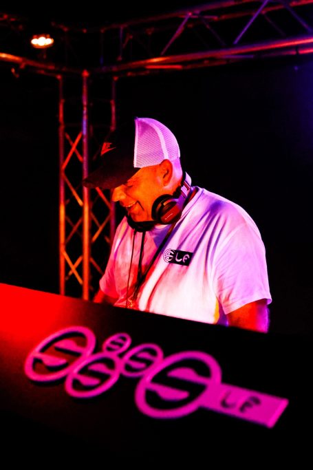 DJ an einem Mischpult mit Kopfhörern, umgeben von buntem Licht und Technik.