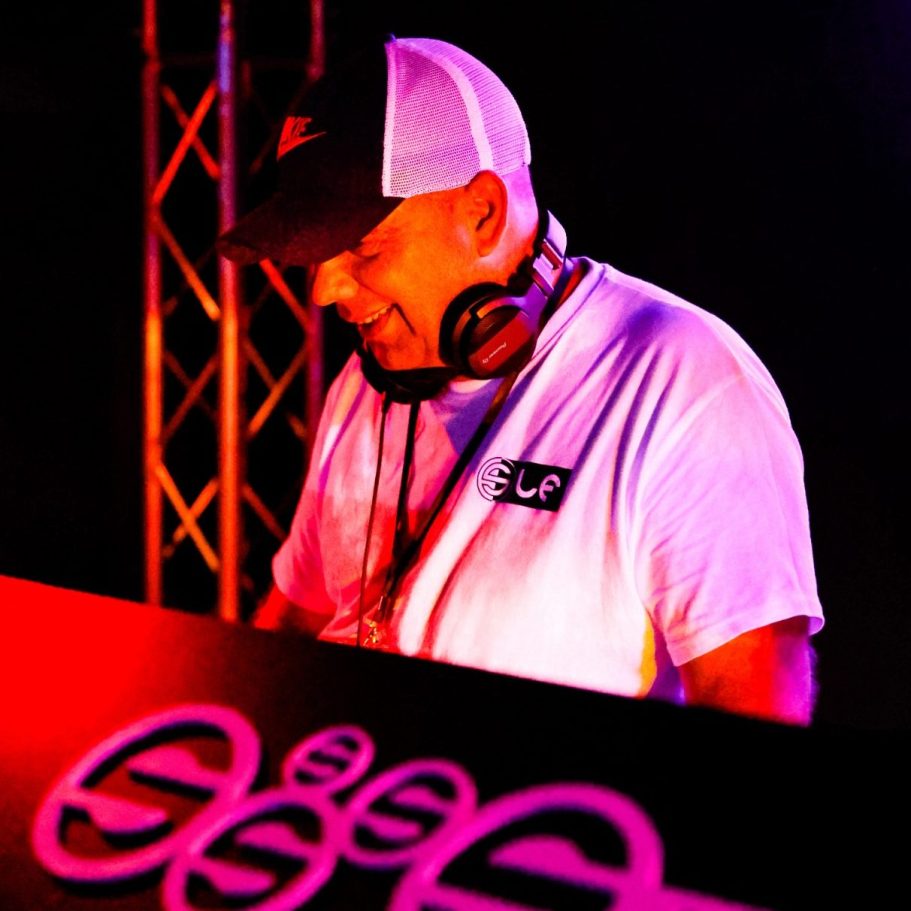 DJ in weißem T-Shirt und Baseballcap, konzentriert am Mischpult. Bunte Lichter im Hintergrund.