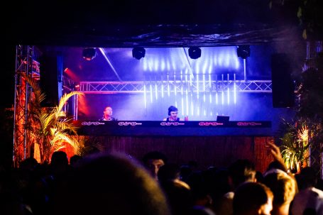 DJ spielt auf einer Bühne mit bunten Lichtern, Publikum tanzt davor.