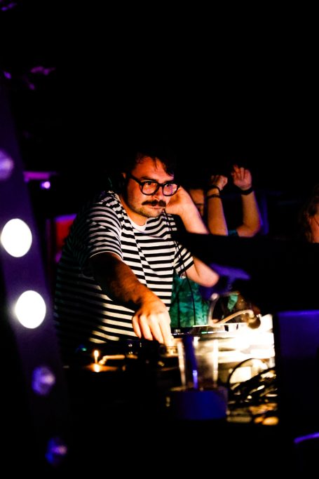 DJ in gestreifter Kleidung an einem Mischpult in einer dunklen, beleuchteten Umgebung.