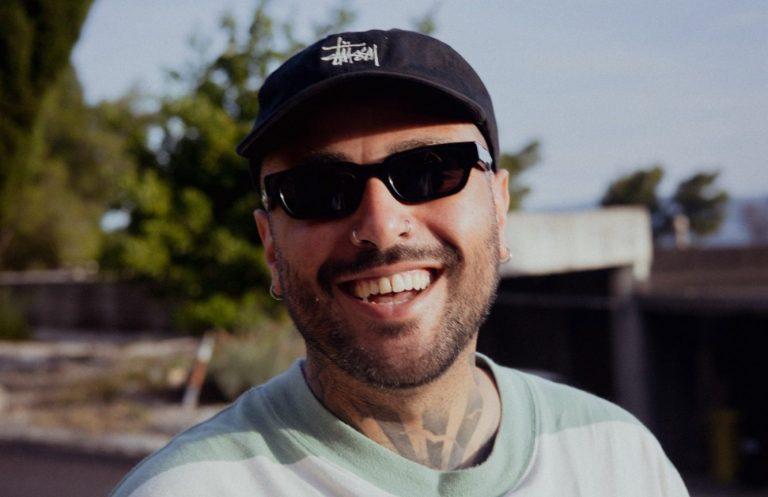 DJ Cookie Lächelnder Mann mit Sonnenbrille und Tattoo, trägt ein gestreiftes Oberteil.
