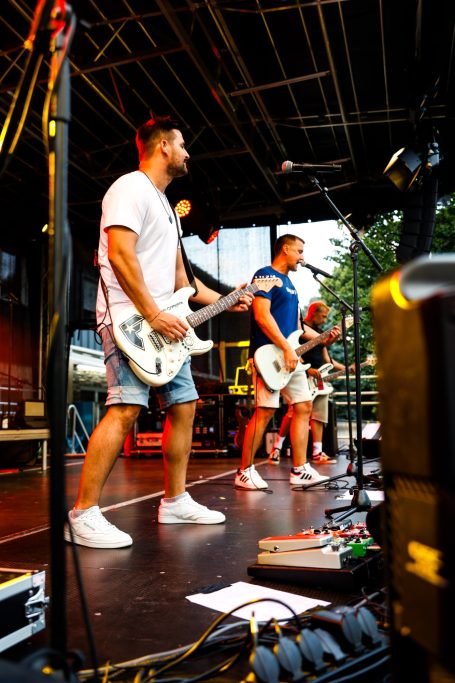 Zwei Musiker auf der Bühne mit Gitarren, im Hintergrund beleuchtete Bühne und Zuschauer.