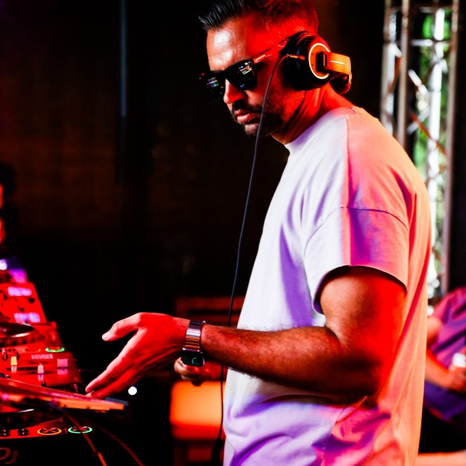 DJ in Sonnenbrille, beim Auflegen an einem Mischpult mit bunten Lichtern im Hintergrund.