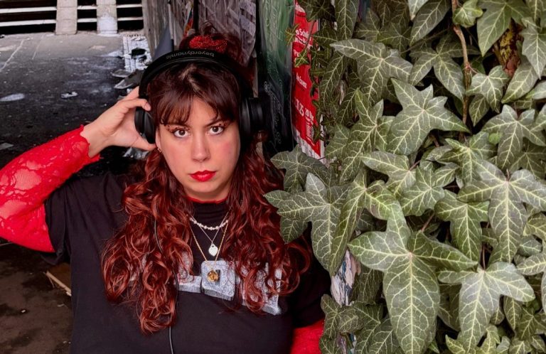 Pöbeln & Pogen Junge Frau mit Kopfhörern, rotem Oberteil und langen, braunen Haaren vor einer Pflanzenwand.