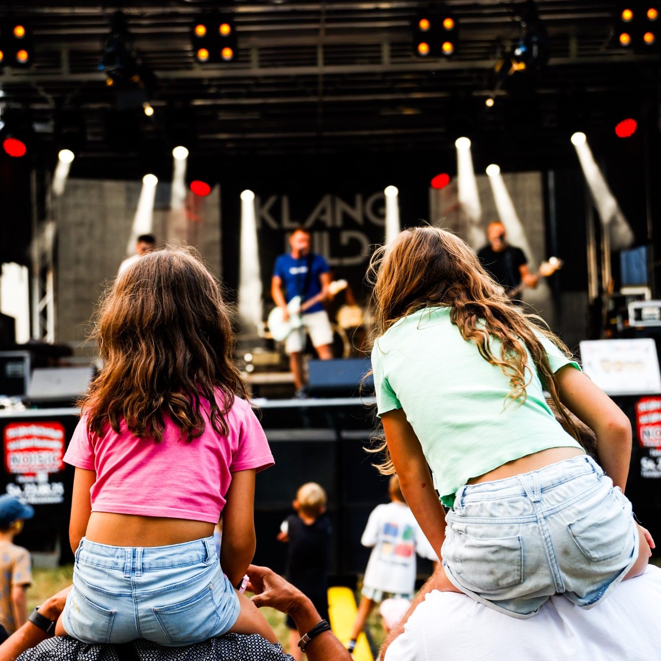 Zwei Kinder auf den Schultern von Erwachsenen schauen einem Konzert mit Band zu.