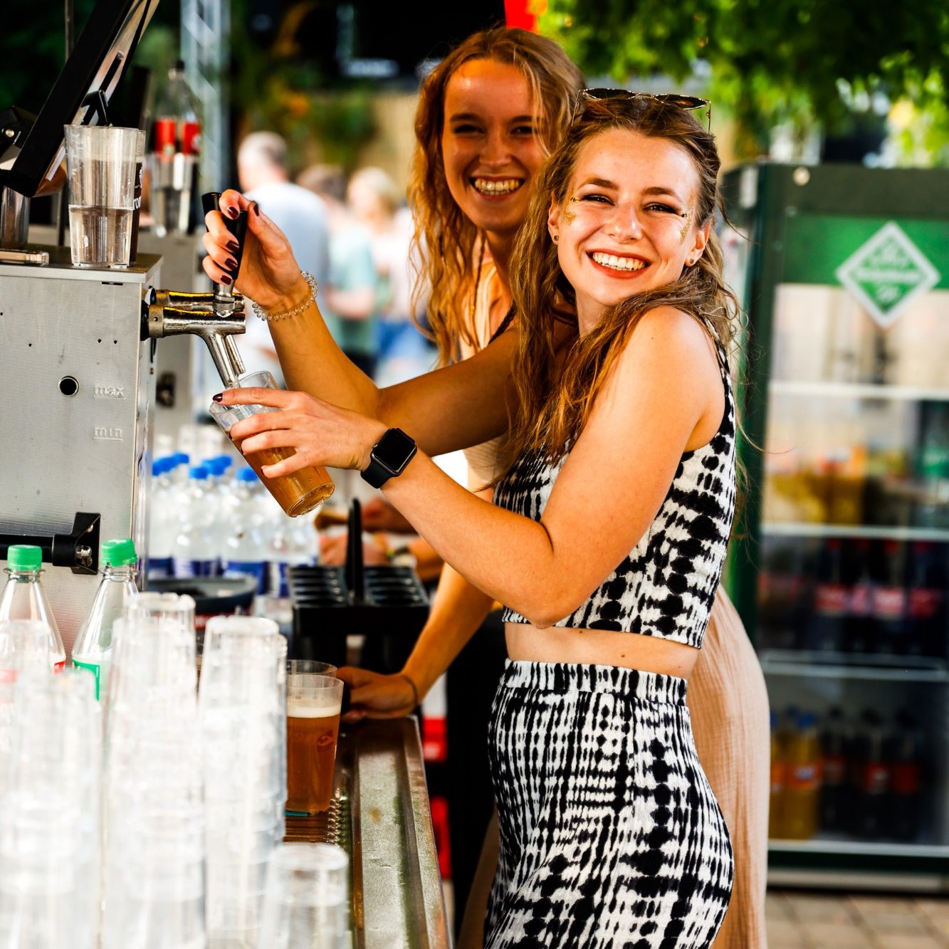 Zwei Frauen lächeln an einem Getränke-Stand, eine hält ein Glas in der Hand.